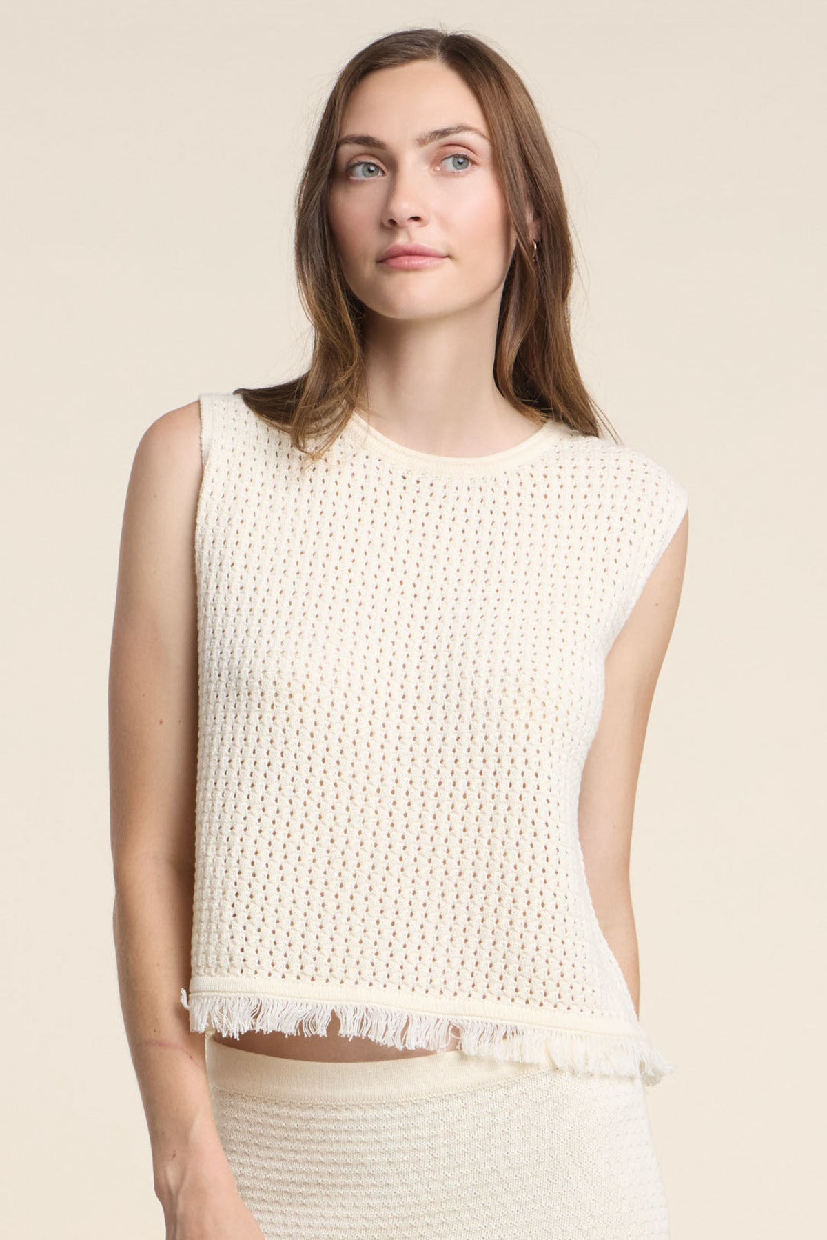 A woman wears the COVA SWEATER by Velvet by Graham & Spencer—a sleeveless, off-white cotton knit top with a fringe hem—standing against a plain beige background.-41374547509441