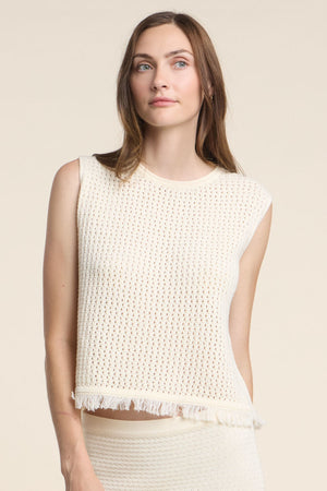 A woman wears the COVA SWEATER by Velvet by Graham & Spencer—a sleeveless, off-white cotton knit top with a fringe hem—standing against a plain beige background.
