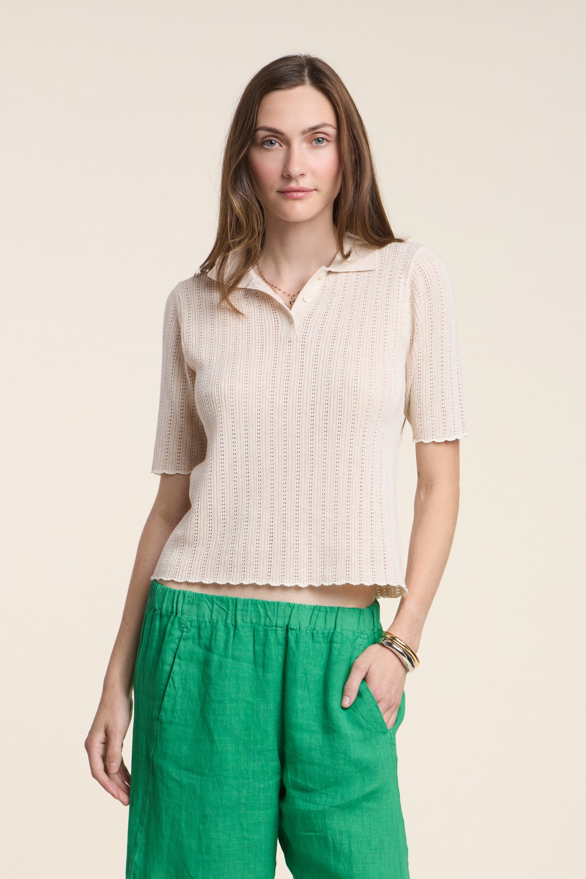 A woman stands against a plain background wearing the DANAE SWEATER POLO by Velvet by Graham & Spencer, featuring scallop detailing, paired with green pants and one hand in her pocket.-41526974021825