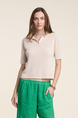 A woman stands against a plain background wearing the DANAE SWEATER POLO by Velvet by Graham & Spencer, featuring scallop detailing, paired with green pants and one hand in her pocket.