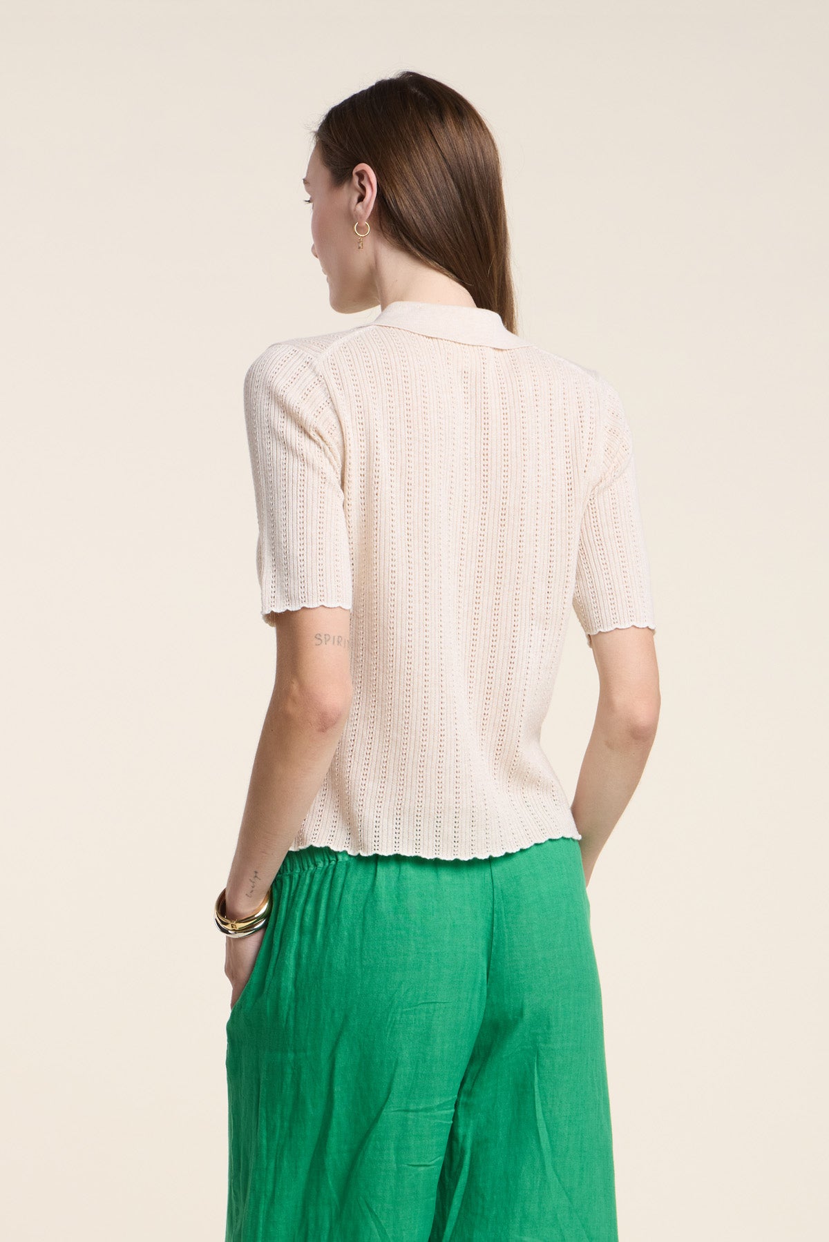 A person stands facing away, wearing Velvet by Graham & Spencer's DANAE SWEATER POLO—a short-sleeve, light beige knit top with subtle pointelle stitch—and green pants against a plain, light background.-41526974087361