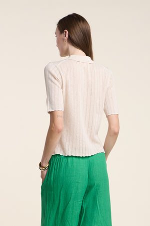 A person stands facing away, wearing Velvet by Graham & Spencer's DANAE SWEATER POLO—a short-sleeve, light beige knit top with subtle pointelle stitch—and green pants against a plain, light background.