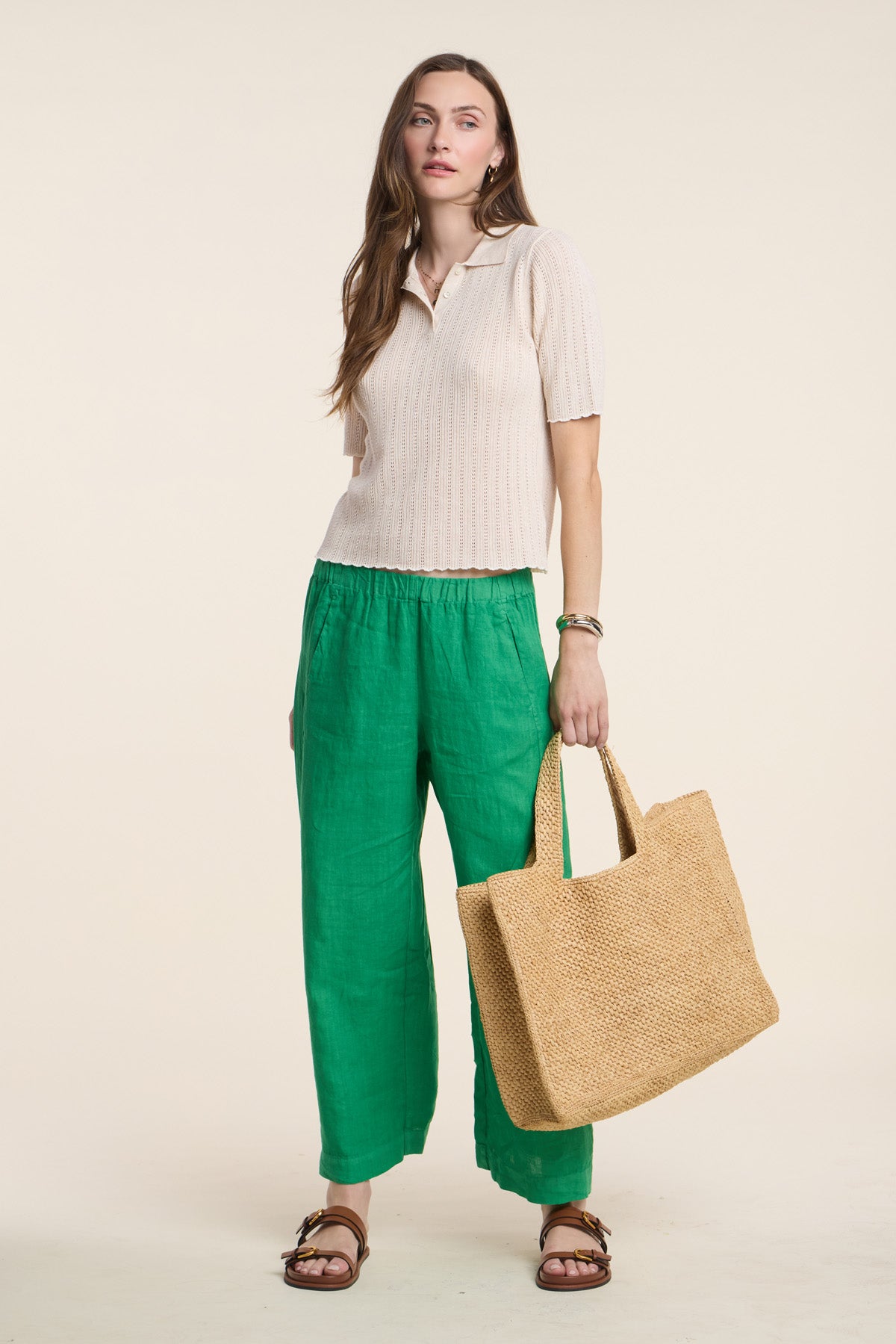 A woman stands against a light background holding a woven tote, wearing the Velvet by Graham & Spencer DANAE SWEATER POLO with short sleeves, paired with green loose pants and brown sandals.