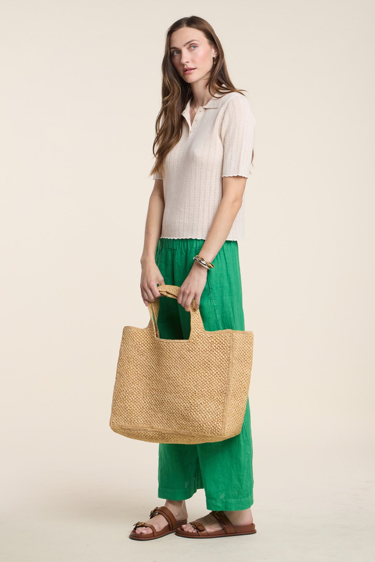 A woman stands against a plain background wearing the DANAE SWEATER POLO by Velvet by Graham & Spencer in beige, styled with green wide-leg pants, brown sandals, and carrying a large woven straw tote bag.-41526974251201