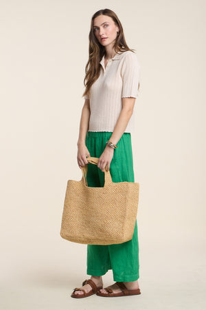 A woman stands against a plain background wearing the DANAE SWEATER POLO by Velvet by Graham & Spencer in beige, styled with green wide-leg pants, brown sandals, and carrying a large woven straw tote bag.