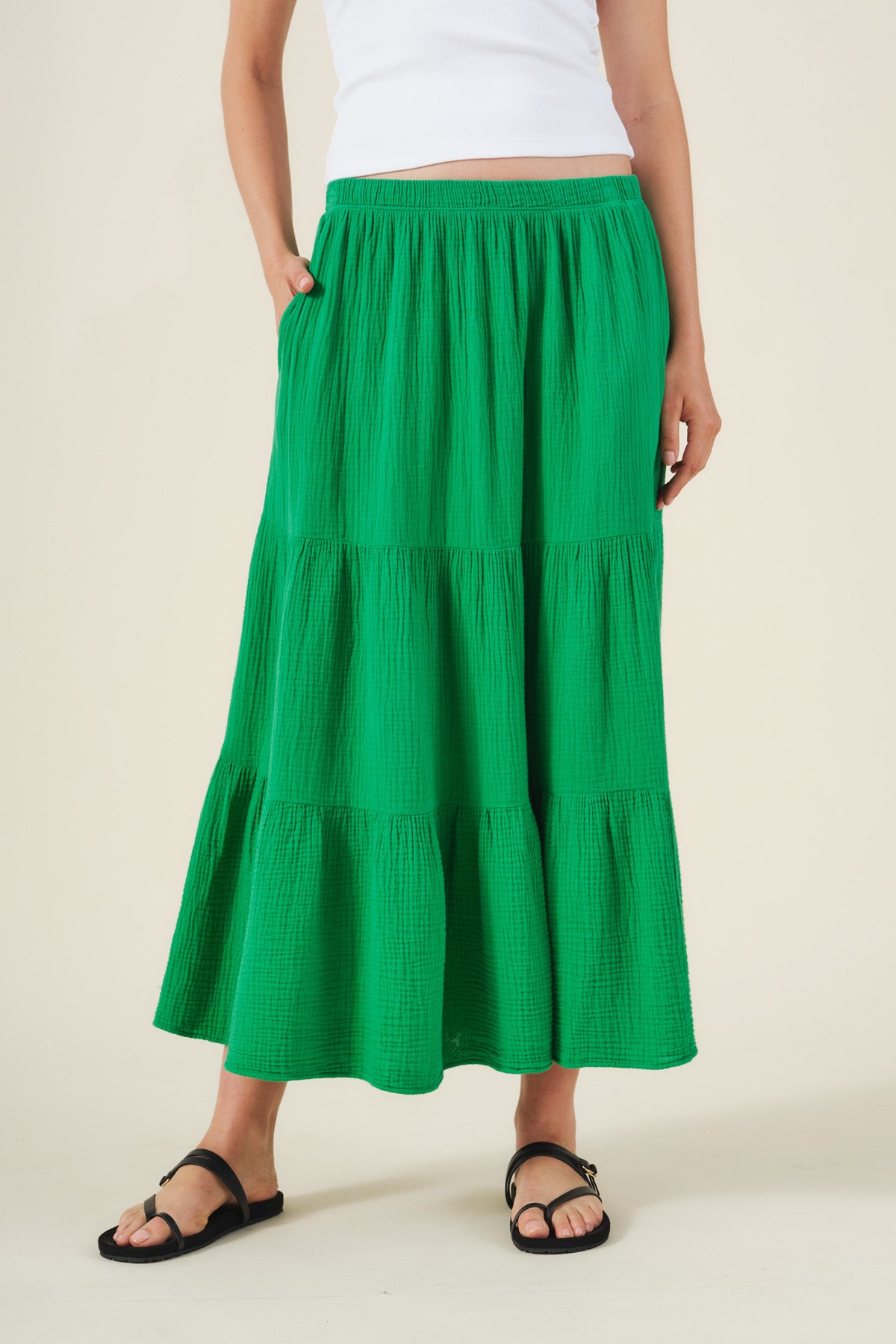 Person wearing a white sleeveless top, the DANIELLE SKIRT by Velvet by Graham & Spencer—a long green cotton gauze skirt with an elastic waist—and black sandals, standing with one hand in the skirt pocket.