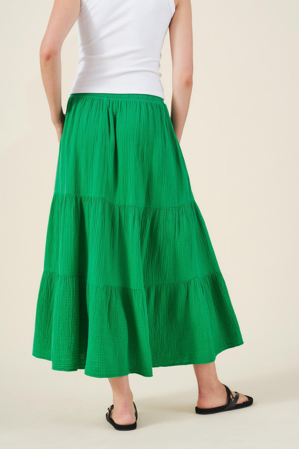 Person wearing a white sleeveless top, Velvet by Graham & Spencer DANIELLE SKIRT in green cotton gauze with an elastic waist, and black sandals, standing against a beige background.-41526997614785