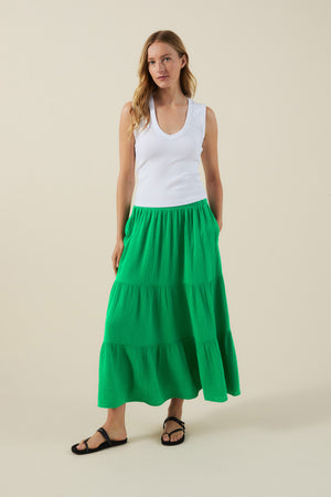 A woman stands against a plain beige background wearing a sleeveless white top, black sandals, and the DANIELLE SKIRT by Velvet by Graham & Spencer—a long green cotton gauze skirt with an elastic waist.