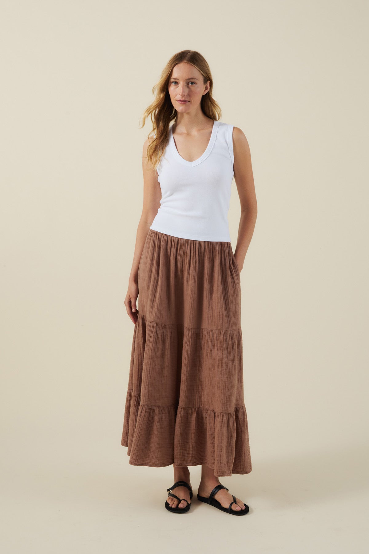 A woman stands against a beige background wearing a white sleeveless top, black sandals, and the DANIELLE SKIRT by Velvet by Graham & Spencer—a brown cotton gauze midi-to-maxi skirt with an elastic waist.