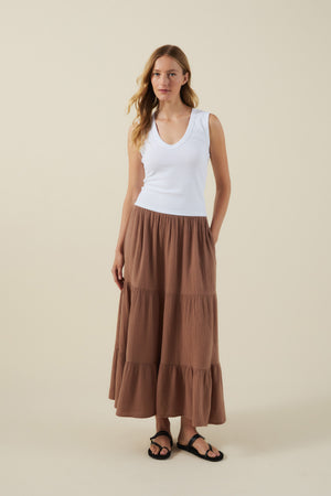 A woman stands against a beige background wearing a white sleeveless top, black sandals, and the DANIELLE SKIRT by Velvet by Graham & Spencer—a brown cotton gauze midi-to-maxi skirt with an elastic waist.