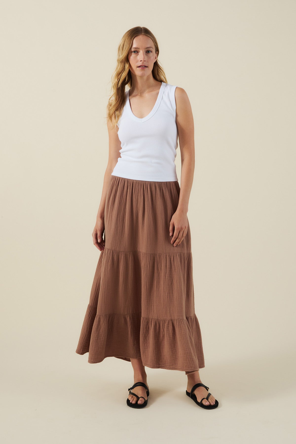 A woman stands against a plain background wearing a white sleeveless V-neck top, black sandals, and the DANIELLE SKIRT by Velvet by Graham & Spencer—a long brown cotton gauze skirt with an elastic waist.