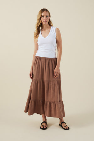 A woman stands against a plain background wearing a white sleeveless V-neck top, black sandals, and the DANIELLE SKIRT by Velvet by Graham & Spencer—a long brown cotton gauze skirt with an elastic waist.