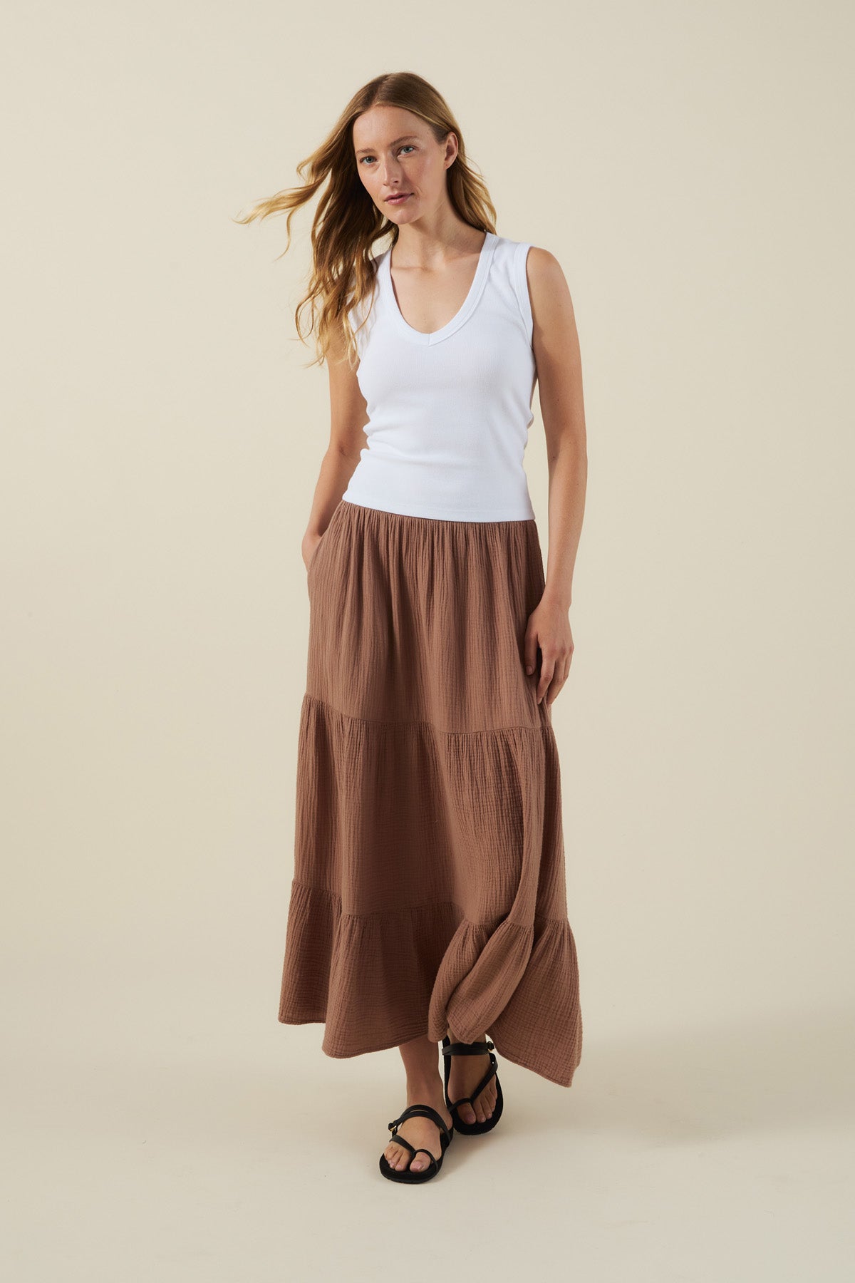 A woman stands against a plain beige background wearing a white sleeveless top, black sandals, and the DANIELLE SKIRT by Velvet by Graham & Spencer—a brown cotton gauze maxi skirt with an elastic waist.
