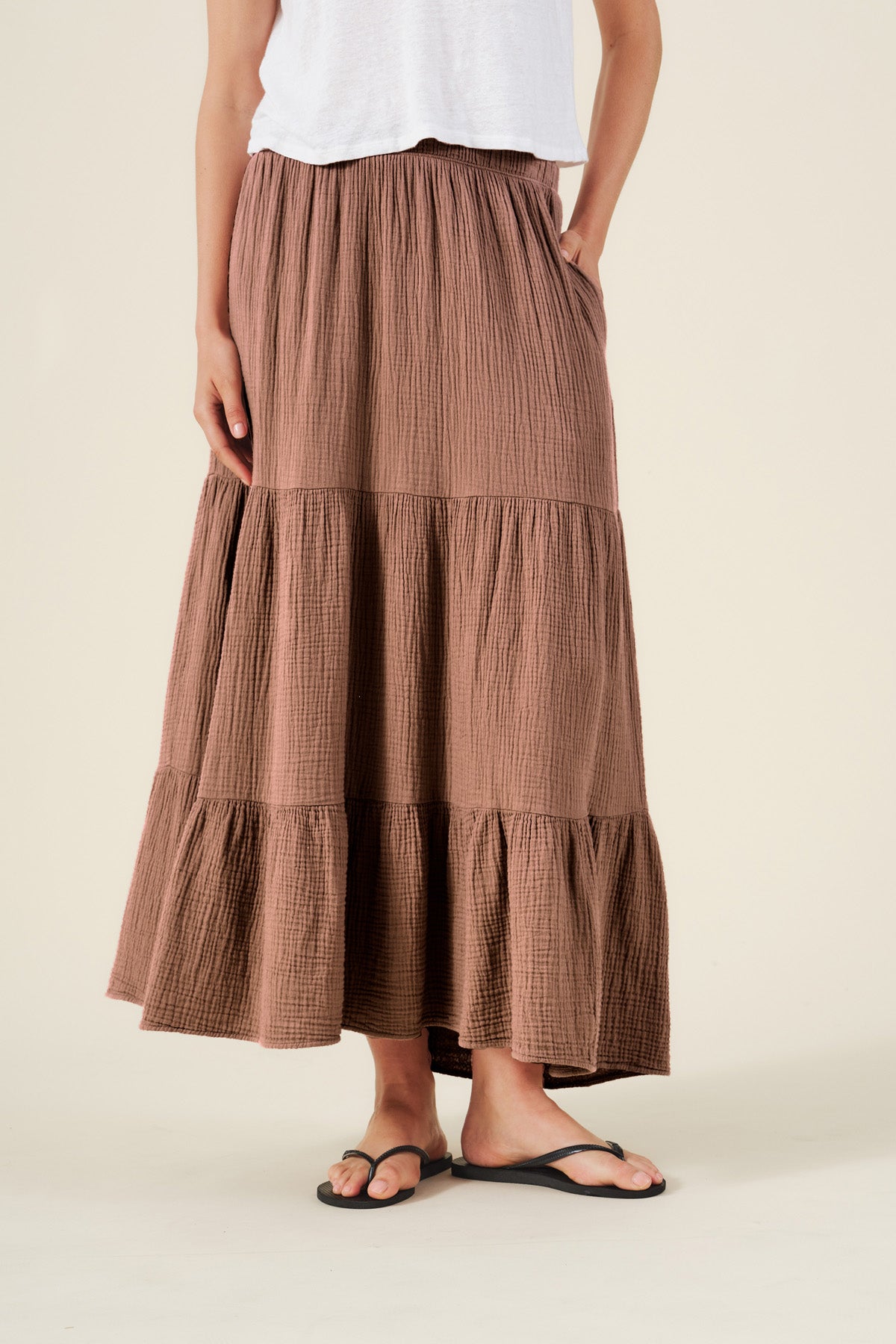 Person wearing a white top, the DANIELLE SKIRT by Velvet by Graham & Spencer—a brown cotton gauze maxi skirt with an elastic waist—and black sandals, standing against a plain background.-41527037722817