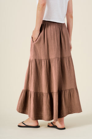 A person wearing a white top, the DANIELLE SKIRT by Velvet by Graham & Spencer—a tiered brown elastic waist skirt—and black flip-flops stands against a plain background.