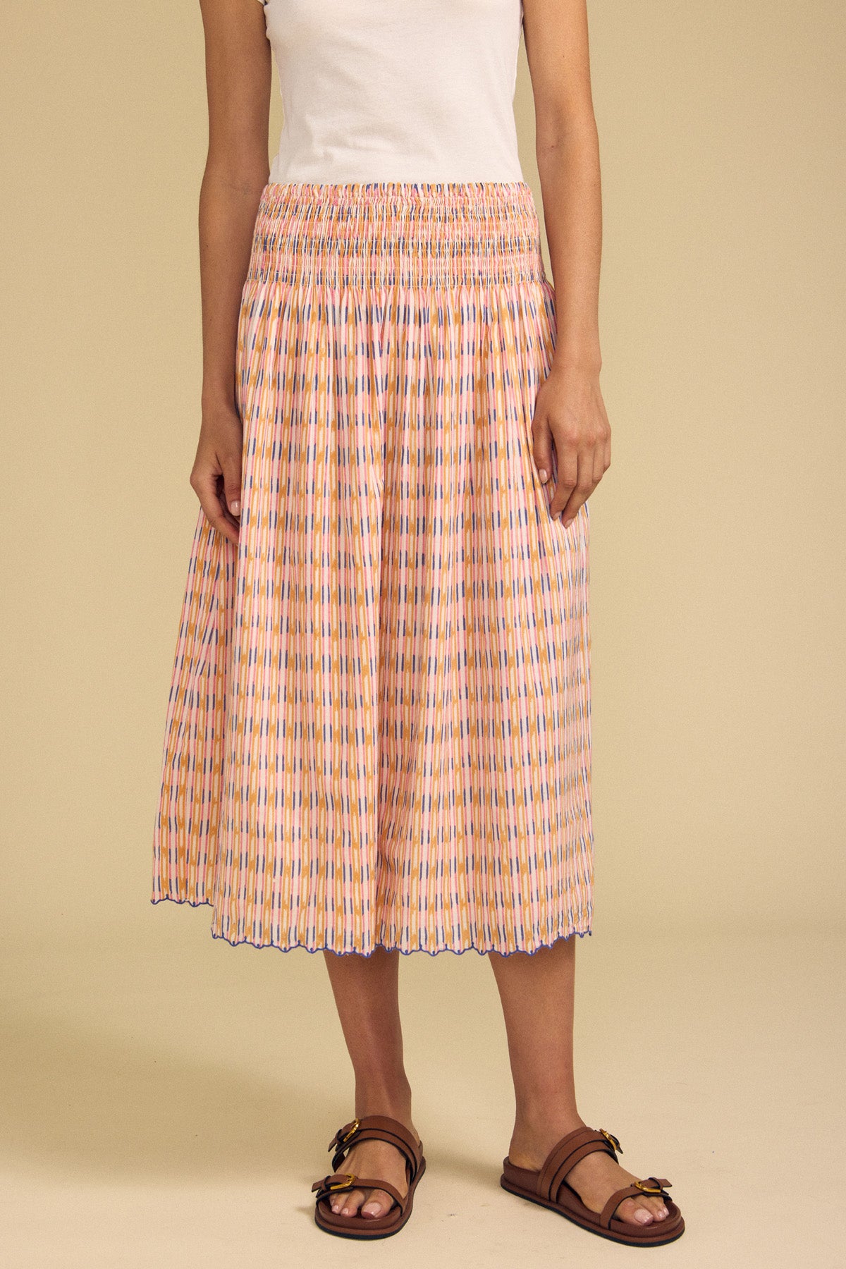 A person wearing a white sleeveless top, the DEBORAH SKIRT by Velvet by Graham & Spencer—a pleated, printed cotton skirt with a smocked elastic waist—and brown sandals stands against a beige background.-41706380099777