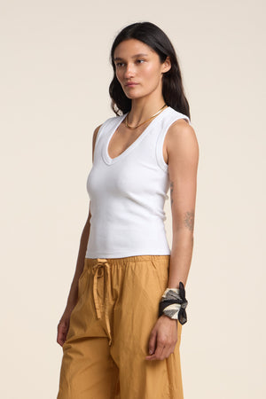 A woman stands against a plain background wearing the DEMI TANK TOP by Velvet by Graham & Spencer, featuring a white ribbed V-neck design, paired with light brown pants. She accessorizes with bracelets on her left wrist and displays a tattoo on her right arm.