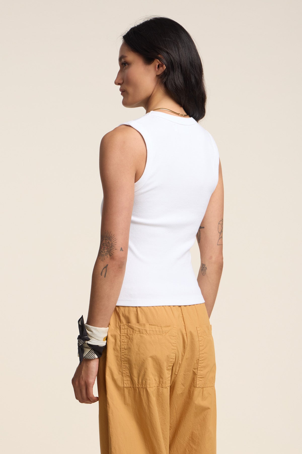 A person with long dark hair, wearing the DEMI TANK TOP by Velvet by Graham & Spencer and tan pants, stands with their back to the camera against a plain light background.