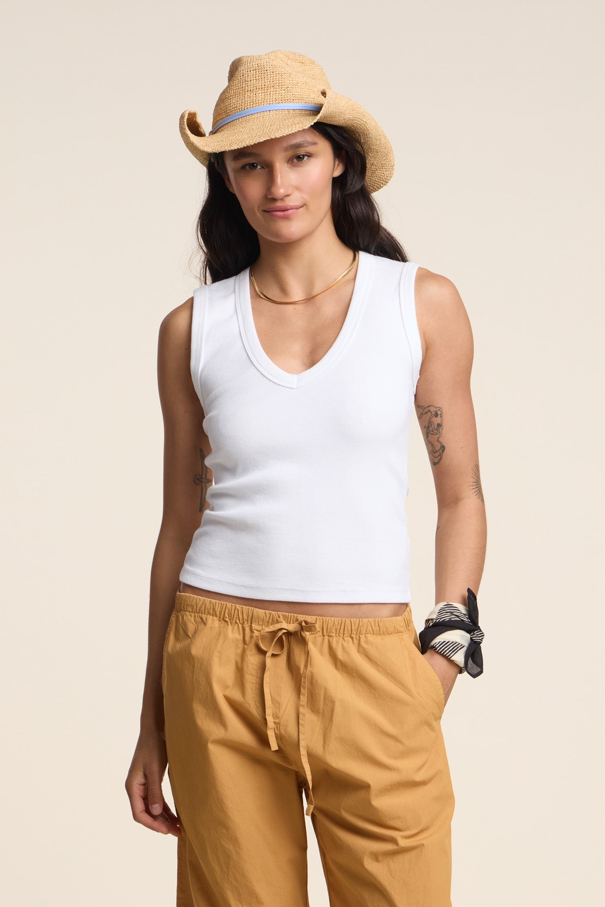 A person wearing a DEMI TANK TOP by Velvet by Graham & Spencer, a straw cowboy hat, and tan drawstring pants stands against a plain background with one hand in their pocket.