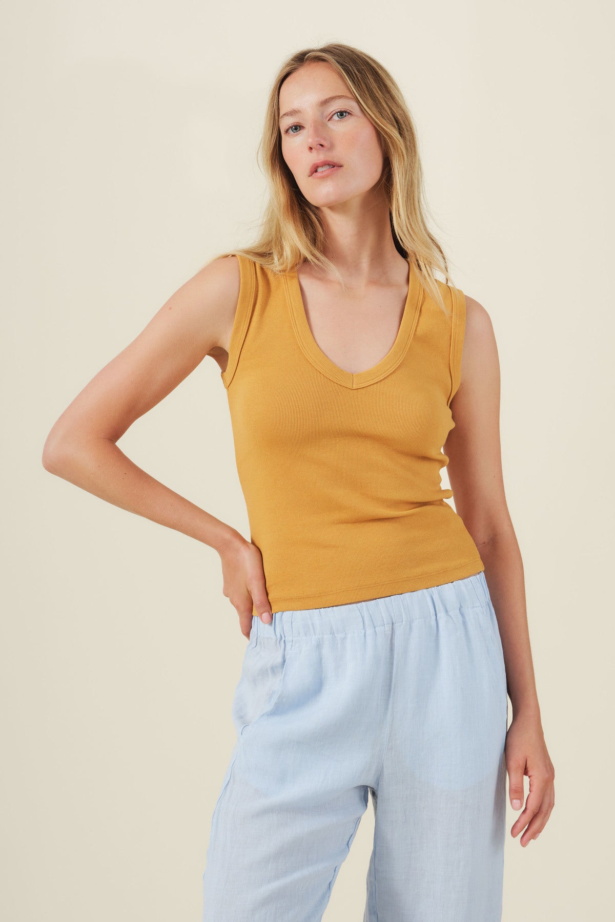 A woman models the DEMI TANK TOP by Velvet by Graham & Spencer, a sleeveless ribbed mustard yellow top, paired with light blue pants against a simple light background.