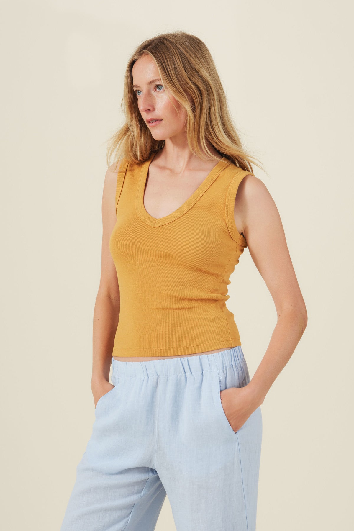 A woman wears the DEMI TANK TOP by Velvet by Graham & Spencer—a mustard yellow ribbed stretch cotton tank with a deep V-neckline—paired with light blue pants, standing hands in pockets against a plain light background. A versatile layering staple.