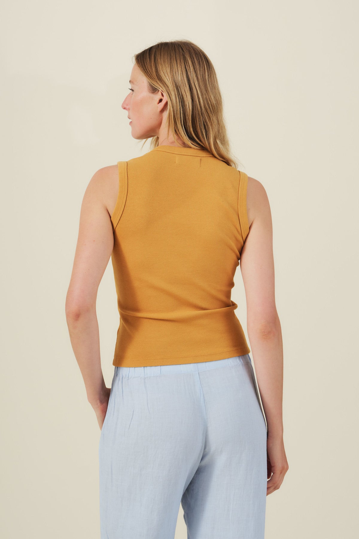 A woman, facing away, wears the DEMI TANK TOP in mustard yellow by Velvet by Graham & Spencer with light blue pants against a plain, light background.-41384926347457