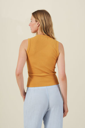 A woman, facing away, wears the DEMI TANK TOP in mustard yellow by Velvet by Graham & Spencer with light blue pants against a plain, light background.