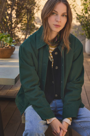 A woman with light brown hair sits on a bench outdoors, wearing the DYLAN BARN JACKET from Velvet by Graham & Spencer, layered over shirts and paired with jeans. Plants and wooden decking are visible in the background.