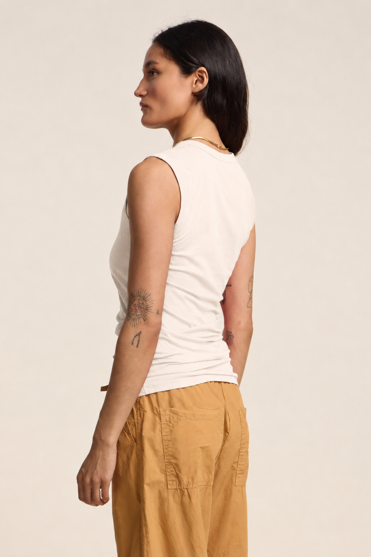 A woman stands sideways against a plain background, showing arm tattoos while wearing the ESTINA TANK TOP by Velvet by Graham & Spencer in white, paired with tan pants.