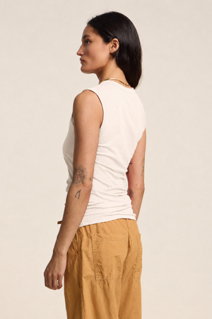 A woman stands sideways against a plain background, showing arm tattoos while wearing the ESTINA TANK TOP by Velvet by Graham & Spencer in white, paired with tan pants.