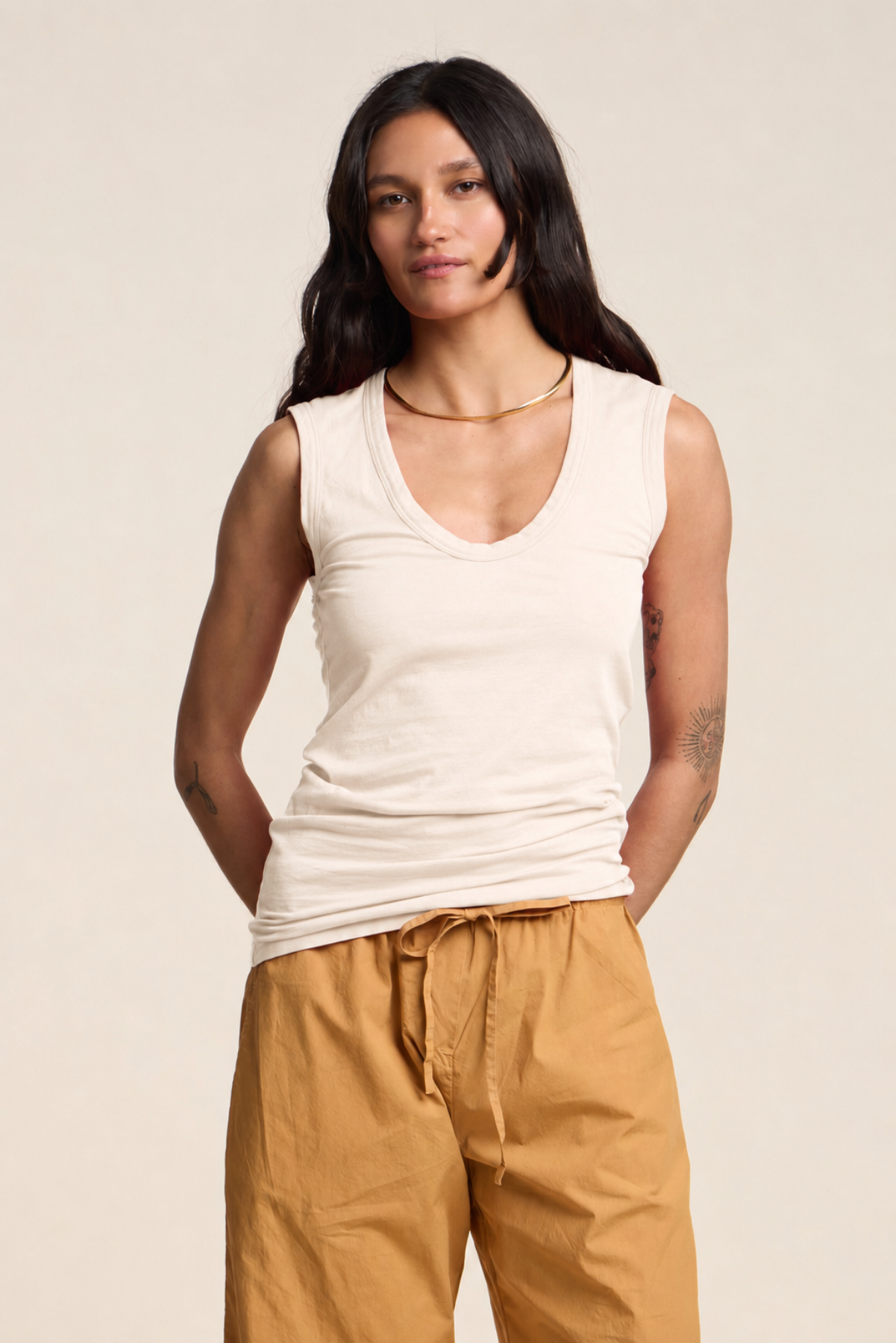 A woman stands against a plain background wearing the Velvet by Graham & Spencer ESTINA TANK TOP in beige, paired with mustard yellow drawstring pants and a gold necklace.