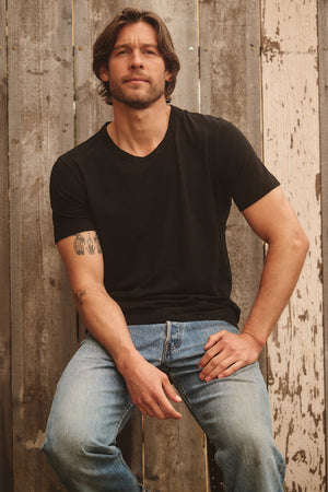 A man with long hair and a beard sits on a stool in front of a wooden fence, wearing the AMARO TEE by Velvet by Graham & Spencer. This black t-shirt, made from soft Peruvian cotton, boasts a classic crew neckline and is an ideal layering piece for any season.