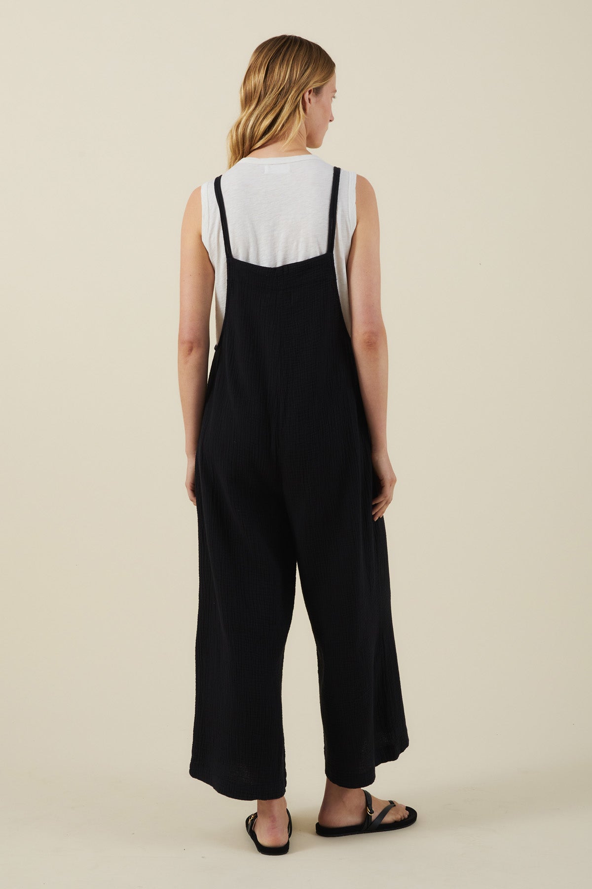 A person with long blonde hair, dressed in a white sleeveless top and the EVERLEE JUMPSUIT by Velvet by Graham & Spencer—featuring wide legs and oversized patch pockets—stands facing away against a plain light background.