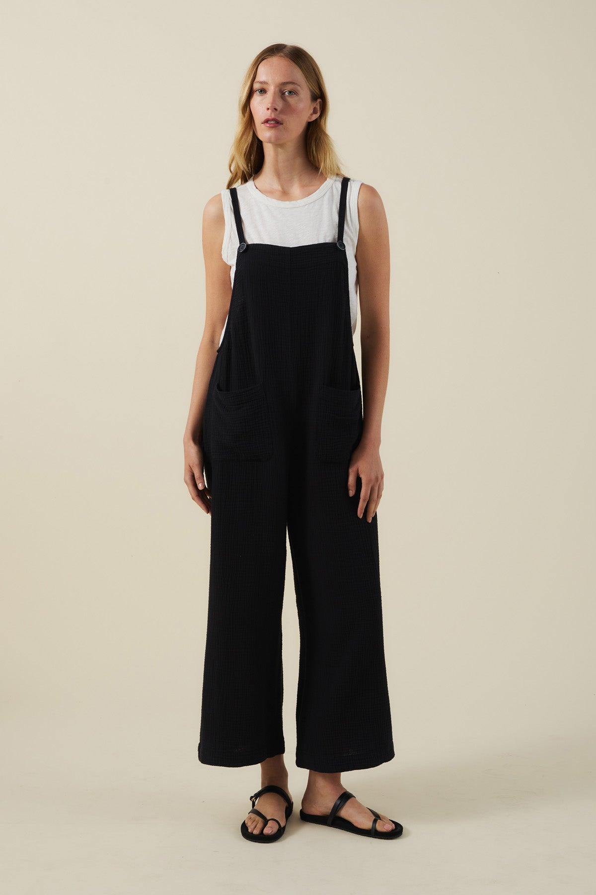 A woman poses against a plain background wearing a white sleeveless top, black sandals, and the EVERLEE JUMPSUIT by Velvet by Graham & Spencer—a black cotton gauze piece with wide legs and oversized patch pockets.-41527322575041