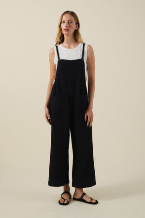 A woman poses against a plain background wearing a white sleeveless top, black sandals, and the EVERLEE JUMPSUIT by Velvet by Graham & Spencer—a black cotton gauze piece with wide legs and oversized patch pockets.