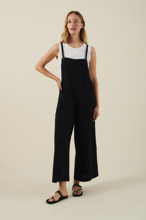 A person stands against a plain background wearing the EVERLEE JUMPSUIT by Velvet by Graham & Spencer—a white, wide-leg cotton gauze jumpsuit with oversized patch pockets—paired with black sandals.