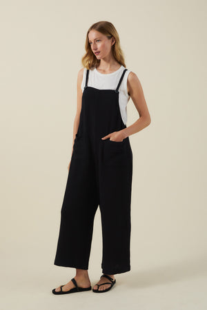 A woman wears the EVERLEE JUMPSUIT by Velvet by Graham & Spencer—a loose black cotton gauze style with wide legs and oversized patch pockets—over a white sleeveless top and black sandals, set against a beige background.