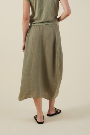 Person stands with their back to the camera, wearing a light olive green t-shirt and the Velvet by Graham & Spencer FAE LINEN SKIRT in an A-line maxi silhouette, paired with black slide sandals against a plain background.