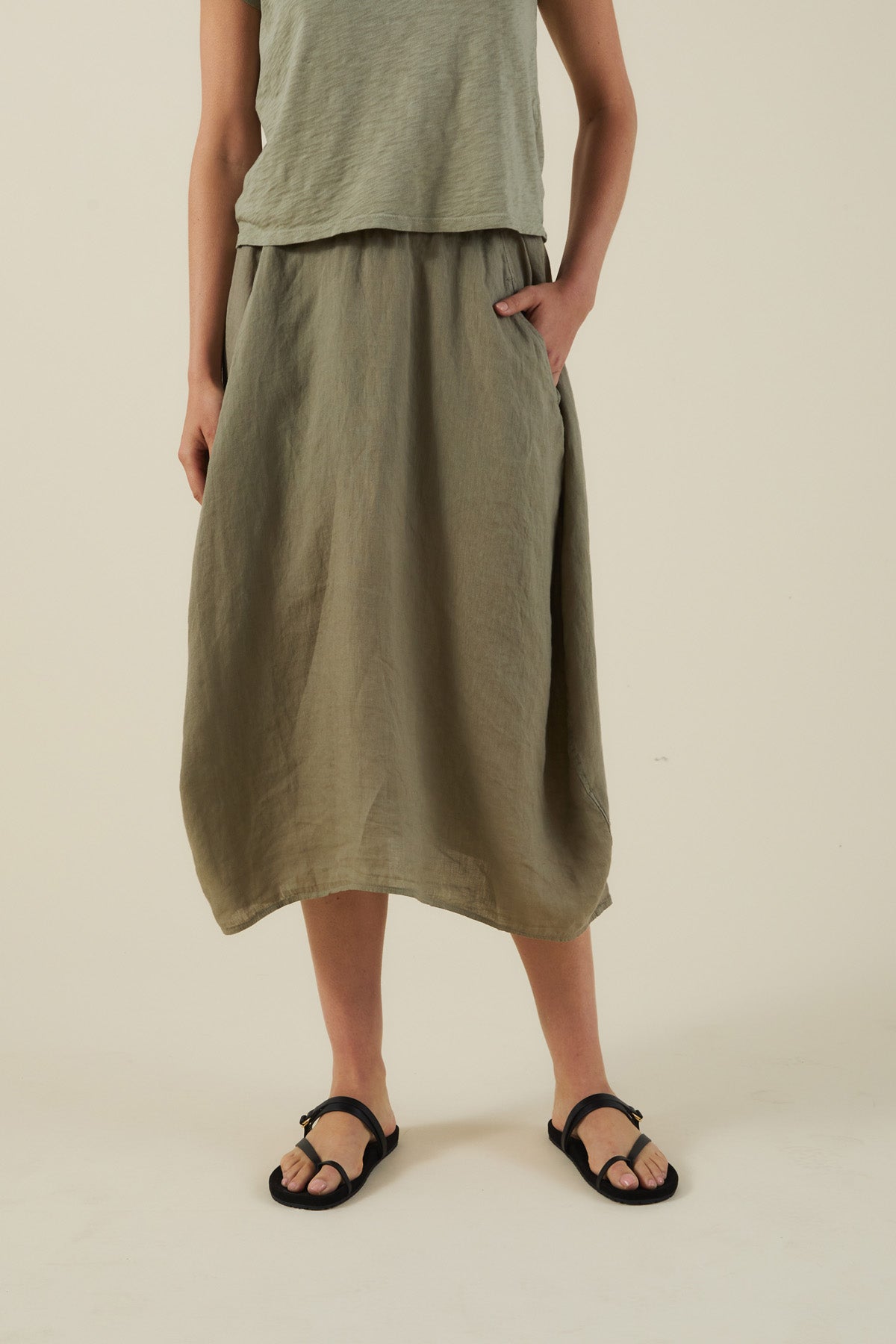 Model wears the Velvet by Graham & Spencer FAE LINEN SKIRT, styled with a loose olive sleeveless top and black sandals, posing with one hand in the pocket against a plain background.-41645028835521