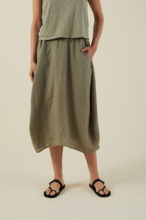 Model wears the Velvet by Graham & Spencer FAE LINEN SKIRT, styled with a loose olive sleeveless top and black sandals, posing with one hand in the pocket against a plain background.