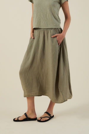 Model wears the FAE LINEN SKIRT by Velvet by Graham & Spencer—an olive green, maxi-length, A-line linen skirt—paired with a loose olive top and black sandals, standing against a plain background with hands in pockets.