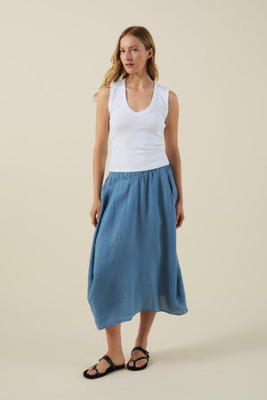 A woman wears the Velvet by Graham & Spencer FAE LINEN SKIRT in blue with an A-line silhouette, paired with a white sleeveless top and black sandals, against a plain light background.