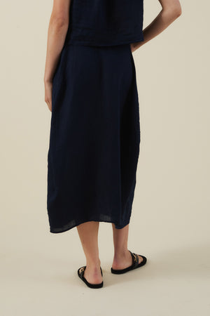 A person stands with their back to the camera, wearing the Velvet by Graham & Spencer FAE LINEN SKIRT in navy blue, paired with a black sleeveless top and black slip-on sandals against a plain beige background.
