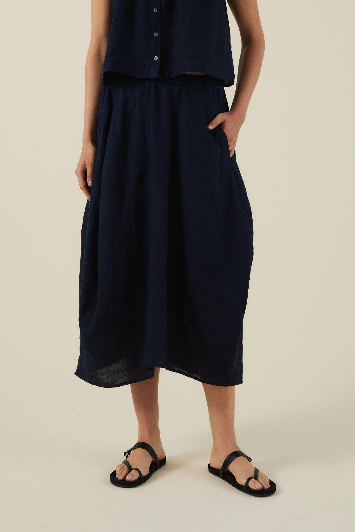A person in a sleeveless navy button-up top, Velvet by Graham & Spencer FAE LINEN SKIRT—navy A-line with pockets—and black sandals stands against a plain background.