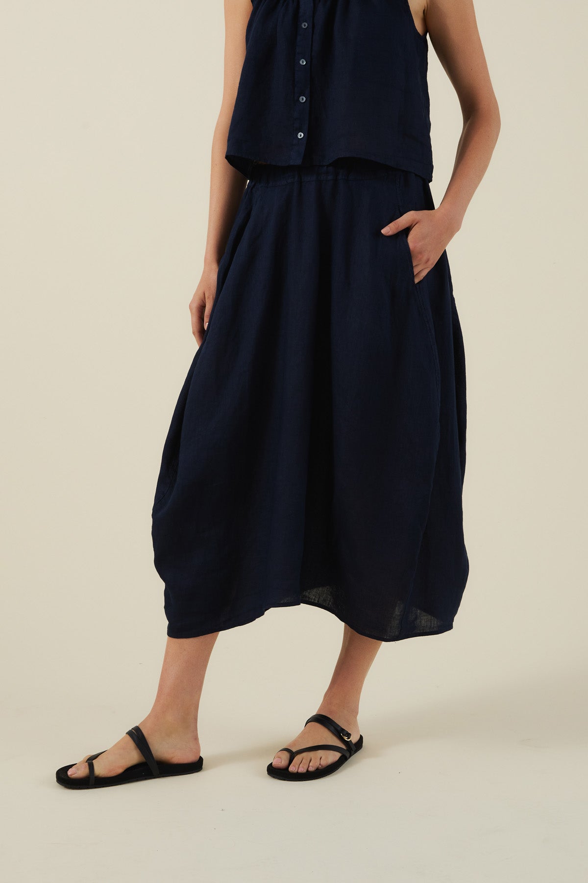 A person wearing a sleeveless navy top and the FAE LINEN SKIRT by Velvet by Graham & Spencer—an A-line, loose linen skirt with pockets—paired with black sandals, stands against a plain light background.