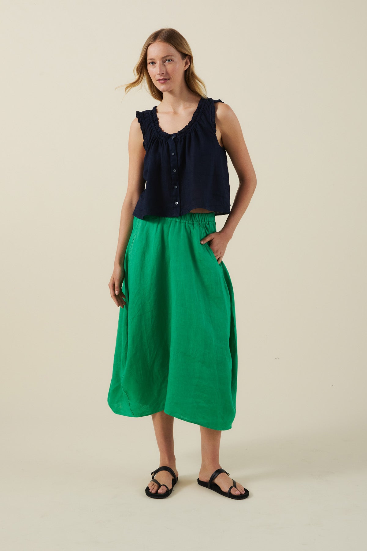A woman stands against a plain background wearing a sleeveless navy top, the FAE LINEN SKIRT by Velvet by Graham & Spencer—an A-line green linen skirt—with black sandals, and one hand in her pocket.-41527342530753