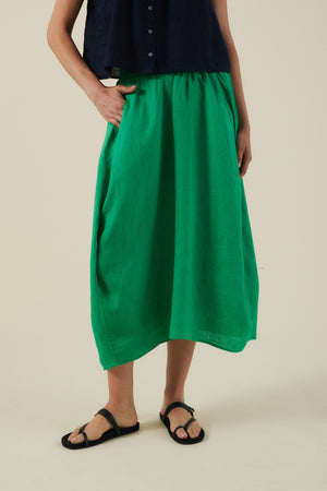 A person wears the Velvet by Graham & Spencer FAE LINEN SKIRT in bright green with an A-line silhouette and pockets, paired with a navy sleeveless top and black sandals, standing against a plain beige background.
