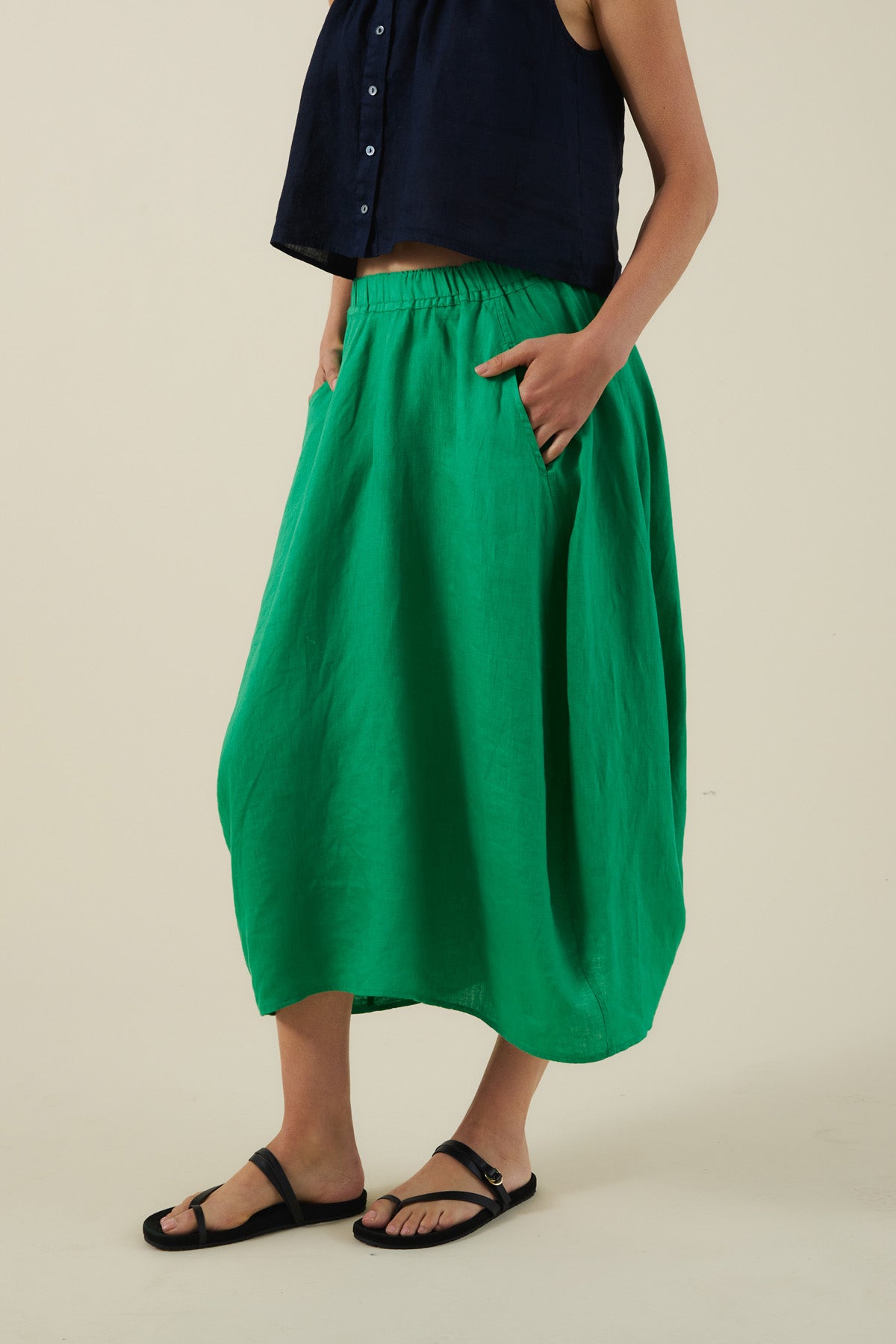 A person wearing a sleeveless navy top, the FAE LINEN SKIRT by Velvet by Graham & Spencer in bright green with an A-line silhouette and pockets, plus black sandals, stands against a plain background.-41527342760129