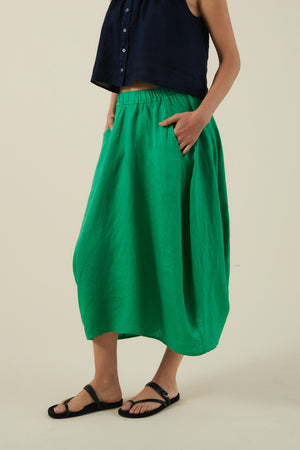 A person wearing a sleeveless navy top, the FAE LINEN SKIRT by Velvet by Graham & Spencer in bright green with an A-line silhouette and pockets, plus black sandals, stands against a plain background.