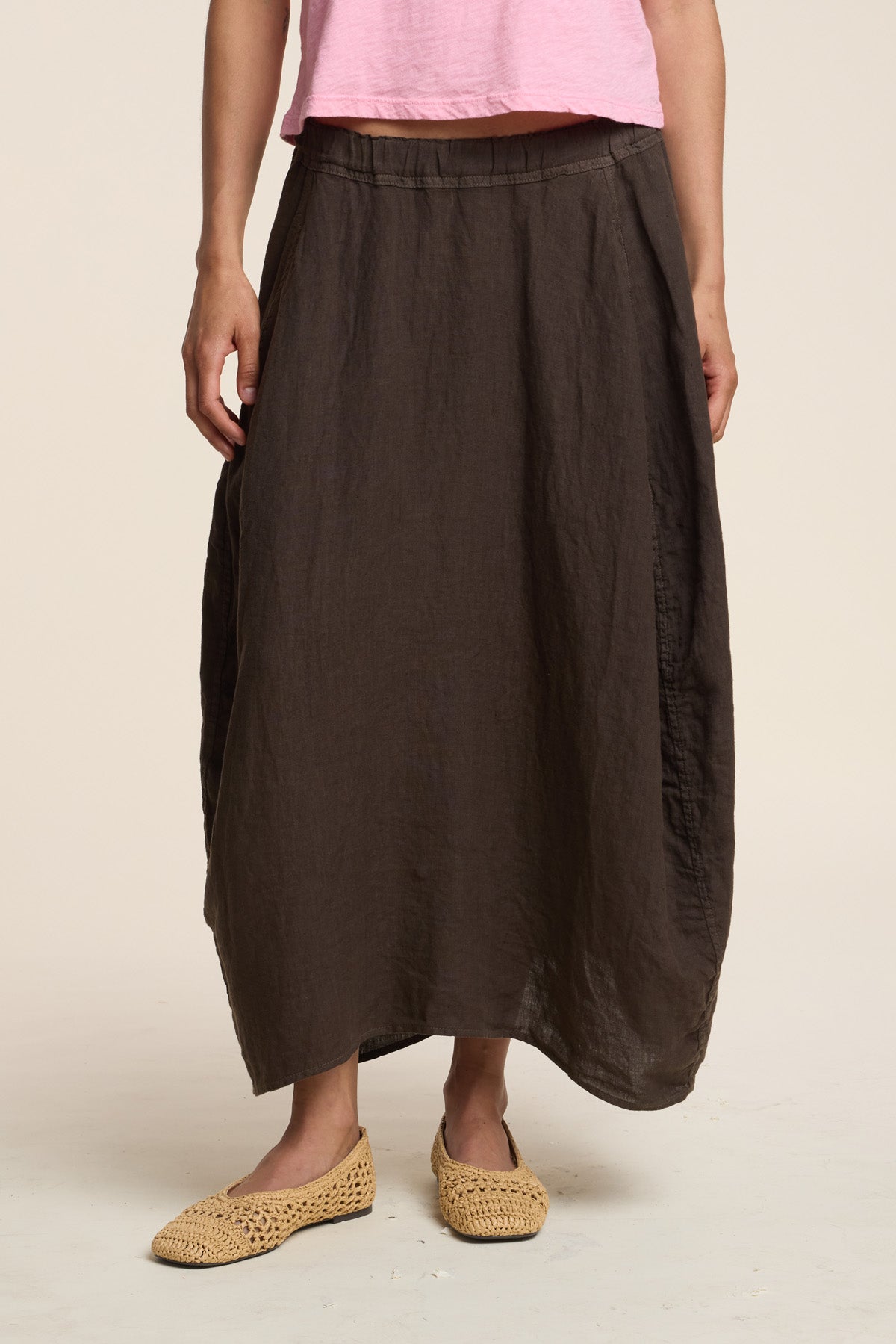 A person wears a pink short-sleeve top, Velvet by Graham & Spencer's FAE LINEN SKIRT in loose brown A-line maxi style, and beige woven shoes, standing against a plain light background.-41460565639361